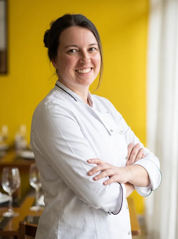 Chef Ana Ferreira, de dólmã branco e braços cruzados, sorrindo no salão do Tempero Fresco